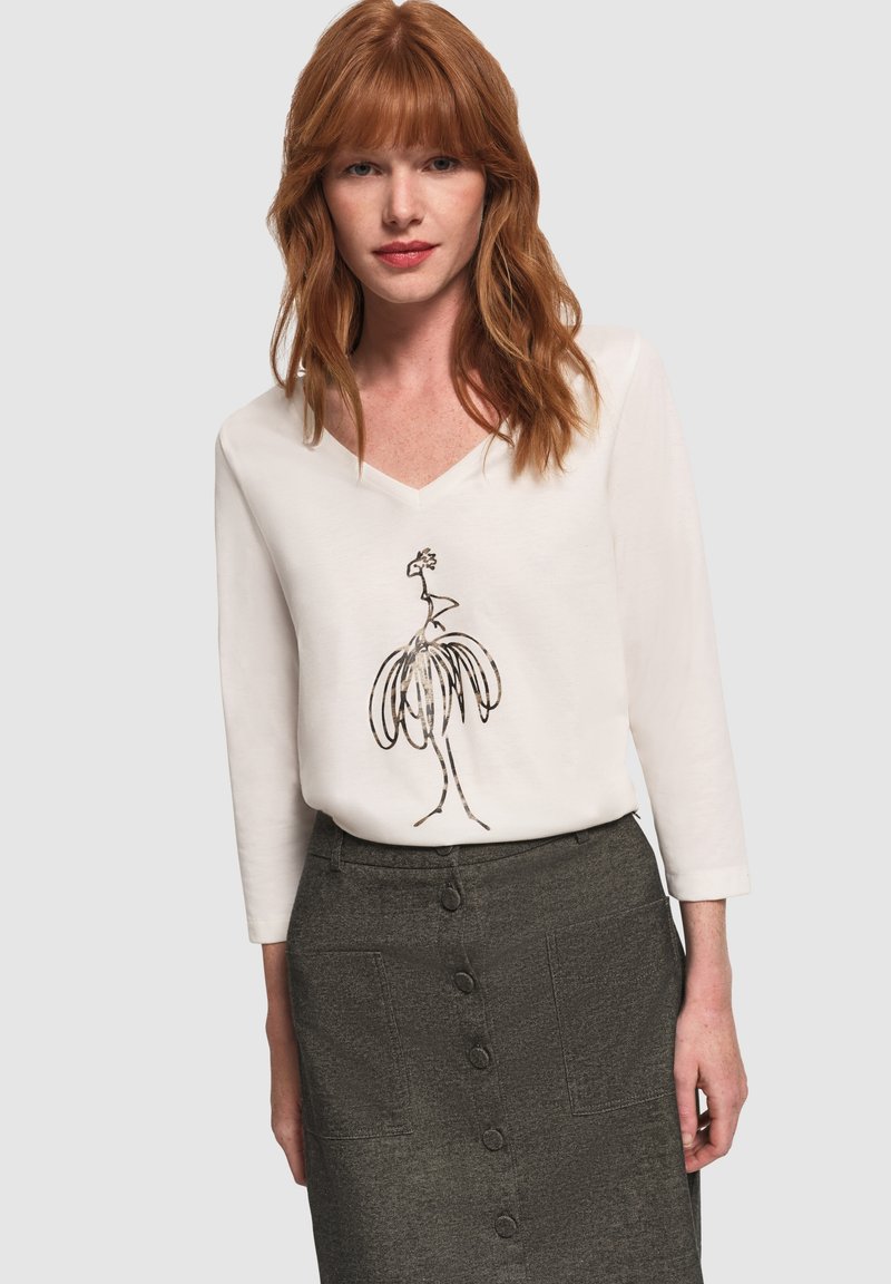 Donna con capelli rossi che indossa un top bianco a scollo a V con un design astratto di uccello in nero e una gonna grigia a vita alta con bottoni.