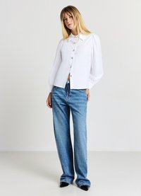 Camicia bianca con colletto a punta, maniche a sbuffo e bottoni dorati. Abbinata a pantaloni in denim blu a vita alta e gamba larga.