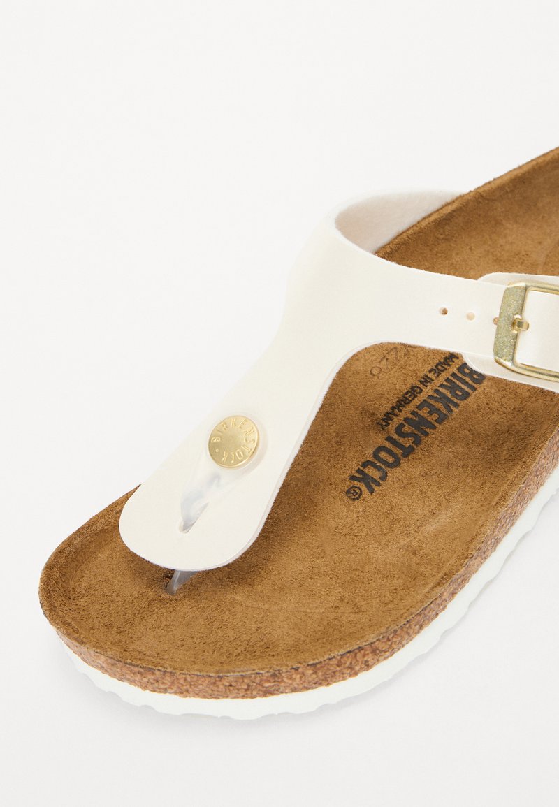 Weiße Zehentrenner-Sandale mit einem strukturierten braunen Fußbett, einem goldenen Schnallenakzent und einer hellen Korksohle. Das Obermaterial ist glatt und flexibel.