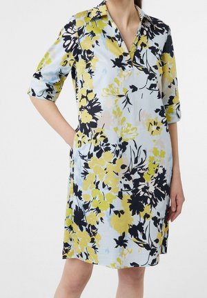 Femme portant une robe à manches courtes avec col, aux motifs floraux jaunes, noirs et bleu clair, debout, une main dans la poche.