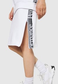 Jupe blanche avec une ouverture zippée latérale et un détail rayé vertical noir et blanc, portée avec des chaussettes blanches et des baskets blanches.