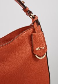Gros plan d’un sac à main en cuir orange texturé avec des bords noirs, une quincaillerie dorée et une étiquette fixée embossée du nom de la marque « abro ».