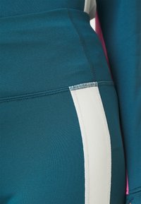 Leggings de sport turquoise en tissu texturé avec une bande blanche sur le côté. Les détails de couture sont visibles.