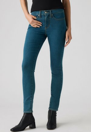 Person iført slim fit blå jeans og sorte ankelstøvler, stående med den ene hånd hvilende på hoften mod en ensfarvet baggrund.