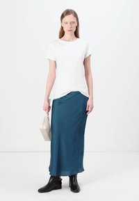 Tricou alb, material ușor, cu guler puțin rotund; fustă maxi din satin teal, textură netedă; botoși negri până la gleznă; ținând o geantă mică albă.