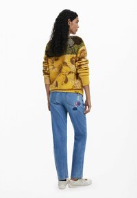 Strickpullover mit einem gelb-braunen Blumenmuster; blaue Jeans mit Blumenstickerei auf der Gesäßtasche; weiße Sneakers.