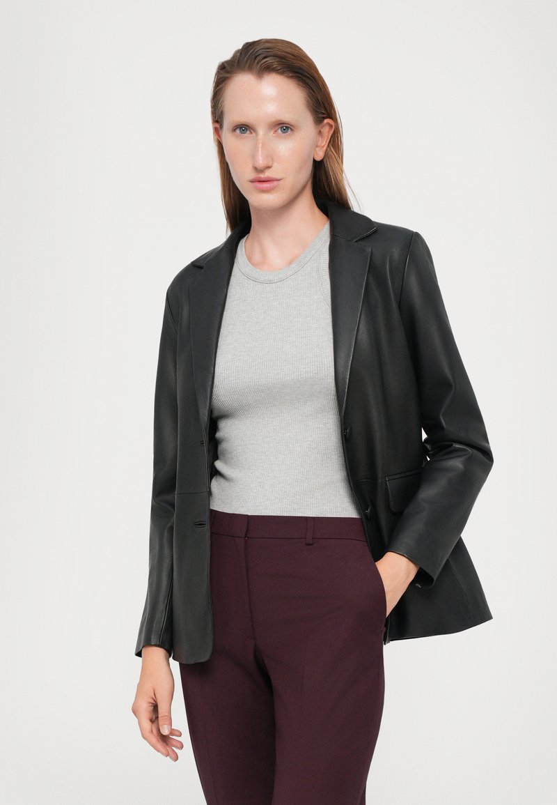 Zwart leren blazer met scherpe revers, gedragen over een grijze ribbeltop. Heeft een getailleerde pasvorm en subtiele voorkruiden, gecombineerd met burgundy broek.