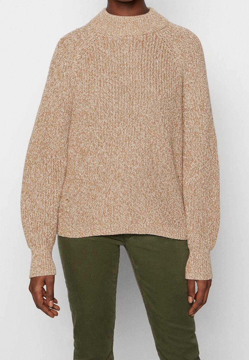 Personne portant un pull en tricot marron clair et blanc avec de longues manches légèrement bouffantes et un pantalon vert olive sur un fond blanc uni.