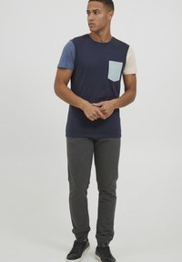 Námořnická modrá tričko s krátkým rukávem v kontrastní modré a světlé béžové, s malou světle modrou kapsou na hrudi; spárováno se šedými tepláky.