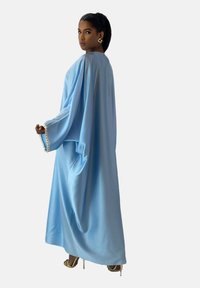 Lyseblå kaftan lavet af glat stof med brede ærmer og perle detaljer ved manchetterne. Modellen bærer guld accessories og høje hæle.