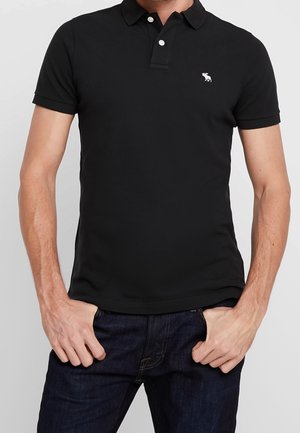 Polo shirt noir à manches courtes, avec un col classique et un petit logo blanc sur la poitrine. Matière en coton lisse.