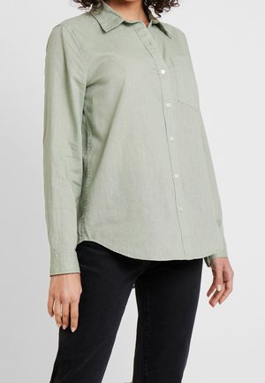 Kobieta ubrana w jasnozieloną koszulę na guziki z długim rękawem i kieszenią na piersi, w połączeniu z czarnymi spodniami, na jednolitym tle.