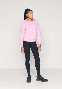 Pink hooded sweatshirt with drawstrings, featuring a front logo, paired with black leggings and black athletic shoes. Smooth fabric textures.