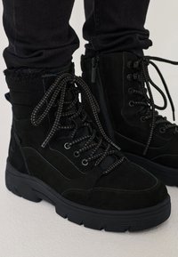 Scarpe in suede nera con lacci per l'inverno, dotate di spesse suole in gomma, indossate con pantaloni neri, mostrando la cerniera laterale e i colli imbottiti.