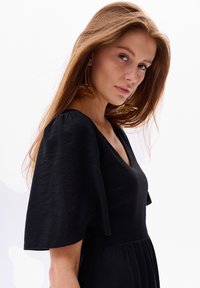 Schwarzes Kleid mit kurzen, schwingenden Ärmeln und tiefem V-Ausschnitt. Glatte Textur und fließendes Design. Ohrringe in Form von goldenen Blättern setzen einen kontrastierenden Akzent.