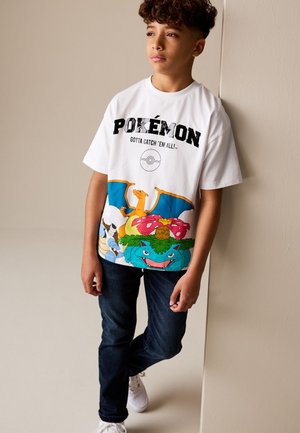 Pois lokkis juustega, valges Pokémoni T-särgis, millel on Charizard, Venusaur ja Blastoise, tumedad teksad ja valged tossud, toetub vastu seina.