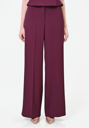 Donna con pantaloni a gamba larga color prugna e tacchi metallici a punta, in piedi su sfondo bianco.