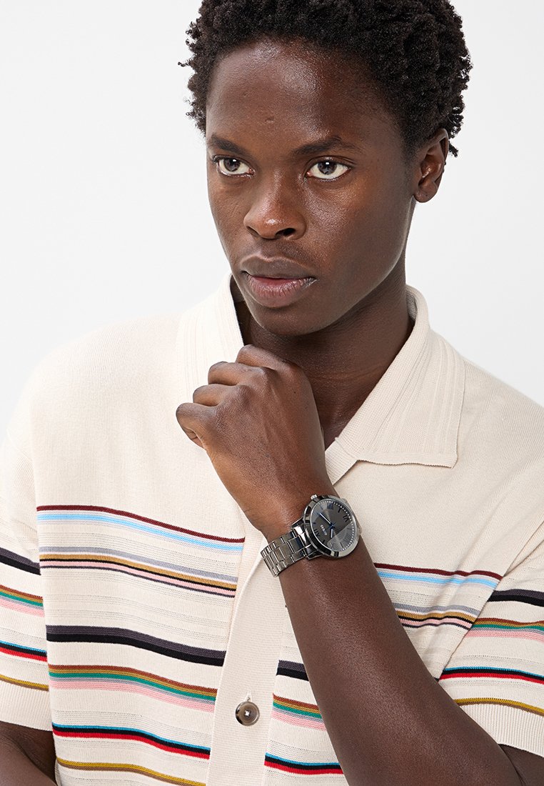 Reloj de pulsera de acero inoxidable con esfera gris oscuro, detalles plateados y correa de eslabones, puesto en la muñeca de una persona que lleva una camisa de rayas.