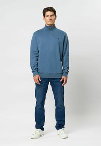 Világoskék bolyhos pulóver fél cipzárral, bordázott mandzsettákkal és aljjal. Sötétkék farmernadrággal és fehér sportcipővel párosítva. Minimális logó részlet.