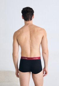 Zwarte katoenen trunks met een bordeauxrode Calvin Klein tailleband, met een gladde textuur en een aansluitend ontwerp, weergegeven vanuit een achteraanzicht.