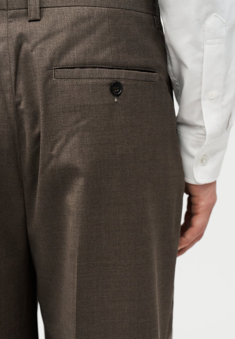 Rückseite brauner Anzughosen mit knöpfbarem Taschen-Detail, getragen mit einem weißen Langarmhemd und sichtbarer Hand.