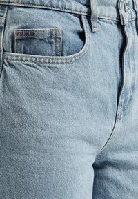 Zbliżenie na jasnoniebieskie dżinsy ukazujące przednią kieszeń, małą kieszeń na monety, szlufkę do paska oraz detale przeszycia.