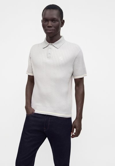 Homme noir portant un polo blanc en maille à manches courtes et un jean bleu foncé, debout avec une main dans la poche sur un fond uni.