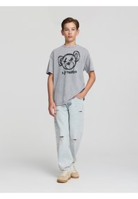 T-shirt grigia oversized con un design grafico nero che presenta il muso di un orso, abbinata a jeans leggeri blu sbiaditi e scarpe da ginnastica beige.