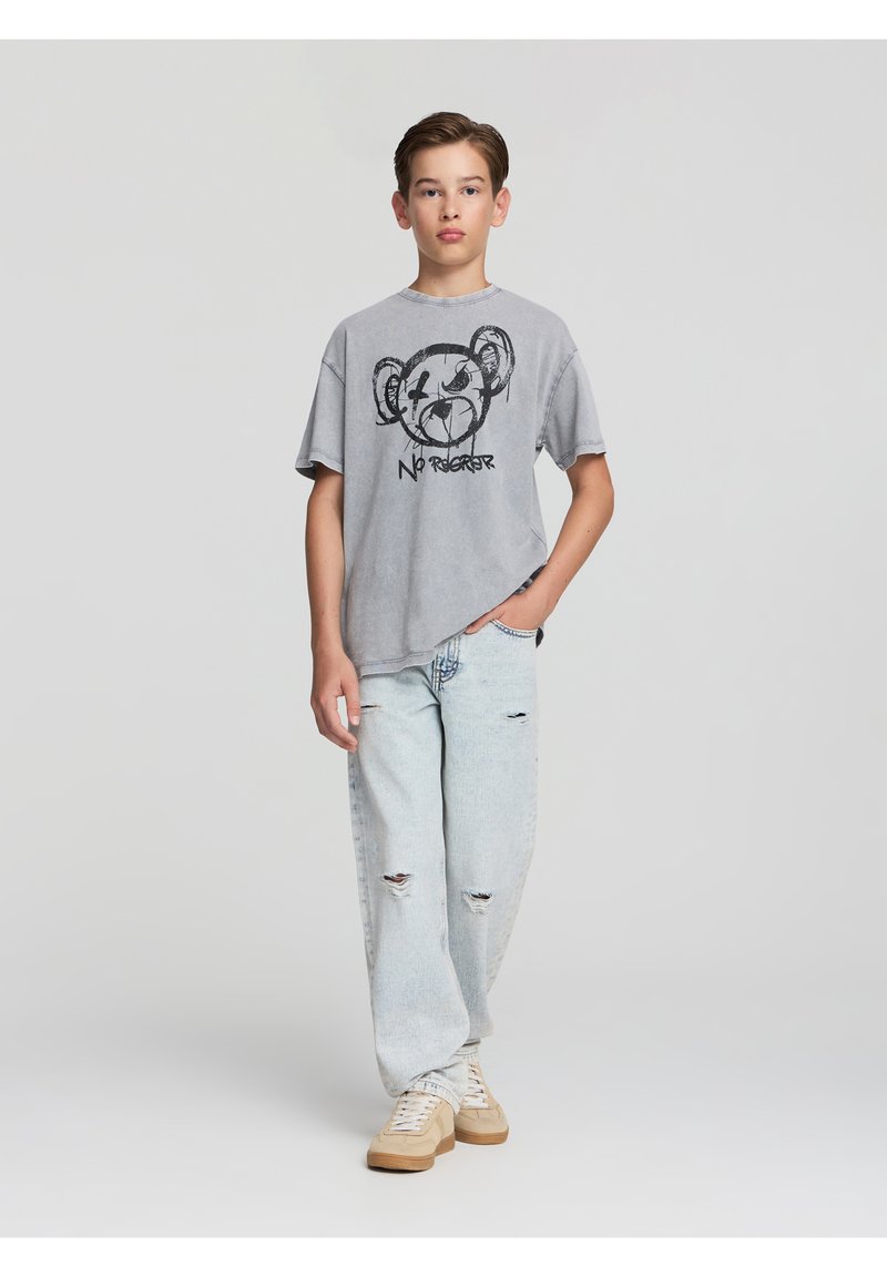 T-shirt grigia oversized con un design grafico nero che presenta il muso di un orso, abbinata a jeans leggeri blu sbiaditi e scarpe da ginnastica beige.
