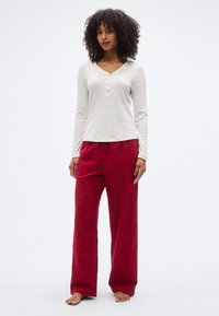 Haut blanc à manches longues avec col en V et trois boutons, associé à un pantalon de pyjama rouge ornée de motifs floraux noirs. Coupe douce et décontractée.