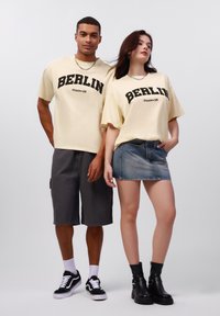 Gule overdimensjonerte t-skjorter med svart "BERLIN" tekst, kombinert med mørke shortser og et denimskjørt, komplementert av svarte ankelstøvler.