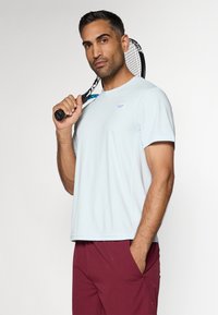 Αθλητική μπλούζα σε ανοιχτό μπλε χρώμα, φτιαγμένη από αναπνεύσιμο ύφασμα, με στρογγυλή λαιμόκοψη και κοντά μανίκια, συνδυασμένη με μπορντό σορτς.