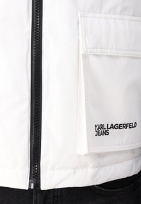 White padded jacket featuring a central black zipper, left chest pocket, and printed "KARL LAGERFELD JEANS" logo in black.