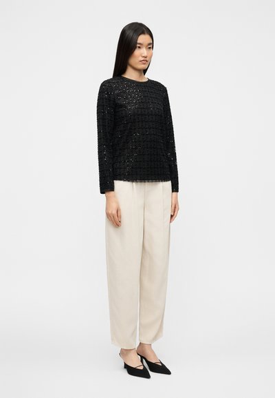 Μαύρο τοπ με μακριά μανίκια και υφασμένο μοτίβο σε πλέγμα· συνδυασμένο με μπεζ παντελόνι φαρδιάς γραμμής και μαύρα ψηλοτάκουνα παπούτσια με μυτερή μύτη, στέκεται σε απλό φόντο.
