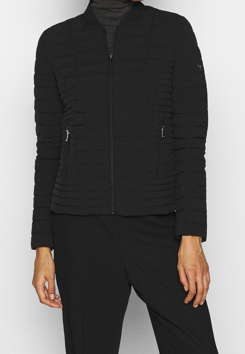 Personne portant une veste matelassée noire zippée sur un col roulé foncé avec un pantalon noir, debout devant un fond blanc uni.