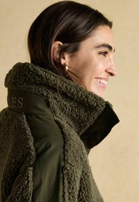 Kurtka oliwkowa z podszewką z polaru, z wysokim kołnierzem, z fakturowaną tkaniną na ramionach i haftowanymi detalami na plecach.