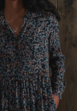 Femme portant une robe florale à manches longues et boutonnée, ornée de petites fleurs bleues et roses, et un collier en argent, se tenant devant un fond en bois.