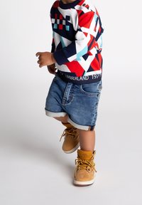 Sudadera de colores con patrones y diseños geométricos, pantalones cortos de mezclilla y botas amarillas hasta el tobillo. El conjunto presenta una cinturilla con el logo de Timberland.