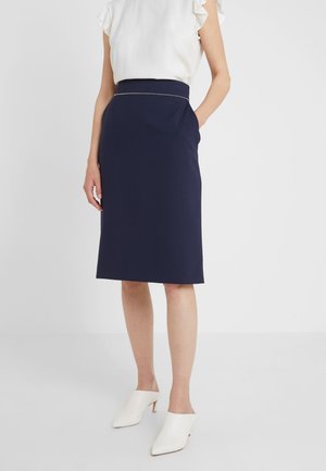 Kobieta ubrana w granatową spódnicę do kolan z bocznymi kieszeniami, dobraną do białej bluzki z falbankami na rękawach oraz białych szpilek z zaostrzonym noskiem.