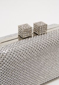 Clutch plateado fabricado con tela texturizada, cubierto de brillantes piedras de estrás. Presenta un cierre rectangular con dos acentos en forma de cubo.