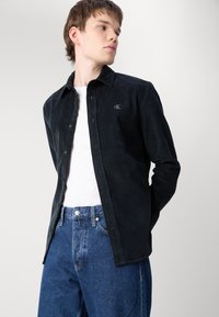 Camicia di velluto a coste nera con bottoni e un piccolo logo, abbinata a jeans denim blu. La camicia presenta un colletto classico e maniche lunghe.