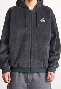 Man i mörkgrå corduroy Adidas zip-up hoodie med ribbade ärmar och midja, matchat med grå byxor.