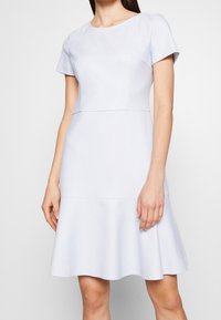 Lichtblauwe jurk met een gladde textuur, korte mouwen, een getailleerde bovenkant en een wijde rok. Heeft een subtiel ontwerpdetail op de borst.