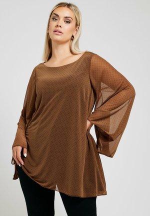 Tunic trasparente marrone con pois neri, maniche ampie e scollatura a barca, abbinata a pantaloni neri aderenti. Tessuto morbido.