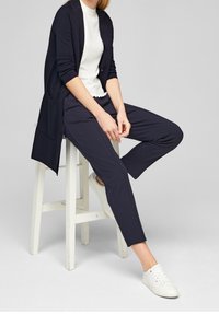 Marinblå lång kofta över en vit topp med vågig nederkant, kombinerad med marinblå byxor och vita sneakers, sittande på en pall mot en grå bakgrund.