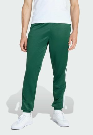 Uomo che indossa pantaloni della tuta verde scuro con strisce laterali bianche e sneakers bianche su uno sfondo chiaro e semplice.