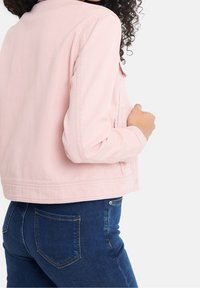 Femme portant une veste en denim rose pâle et un jean bleu foncé, vue de dos sur un fond blanc.