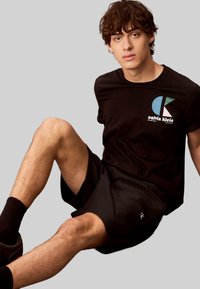 Schwarzes Baumwoll-T-Shirt mit einem grafischen Logo, kombiniert mit schwarzen Shorts, sitzende Position, vor einem grauen Hintergrund. Strukturierte Stoffdetails sichtbar.