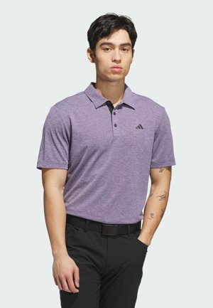 Ung mand med kort mørkt hår iført en lilla Adidas polo shirt og sorte bukser med tatoveringer på venstre arm mod en ensfarvet baggrund.