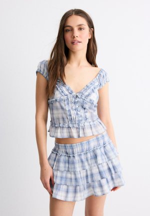 Mladá žena nosí modro-biely kockovaný crop top s volánikmi a zodpovedajúcu viacvrstvovú mini sukňu, stojaca na bielom pozadí.